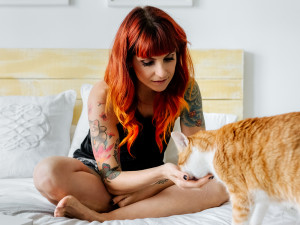 Young modern woman with tattoos and orange hair sitting cross-legged on her bed with her cat 