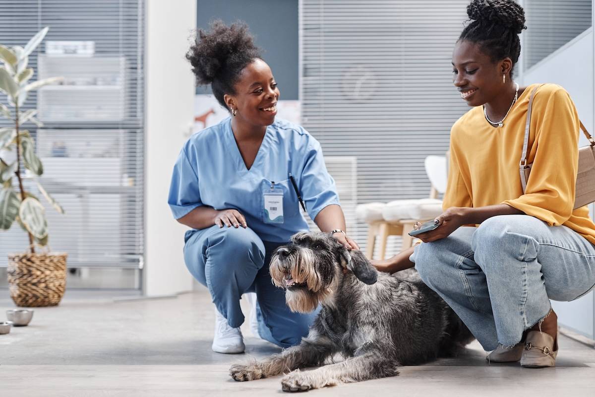 Two women, one in scrubs, crouch down next to a fluffy grey dog