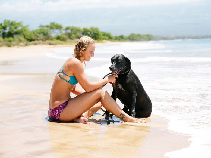 Blonde woman in a teal and purple bikini sitting on the seashore petting her black Labrador dog