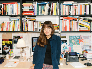 Emma Allen wearing glasses and blue jacket standing in front of her desk with full bookshelves above it