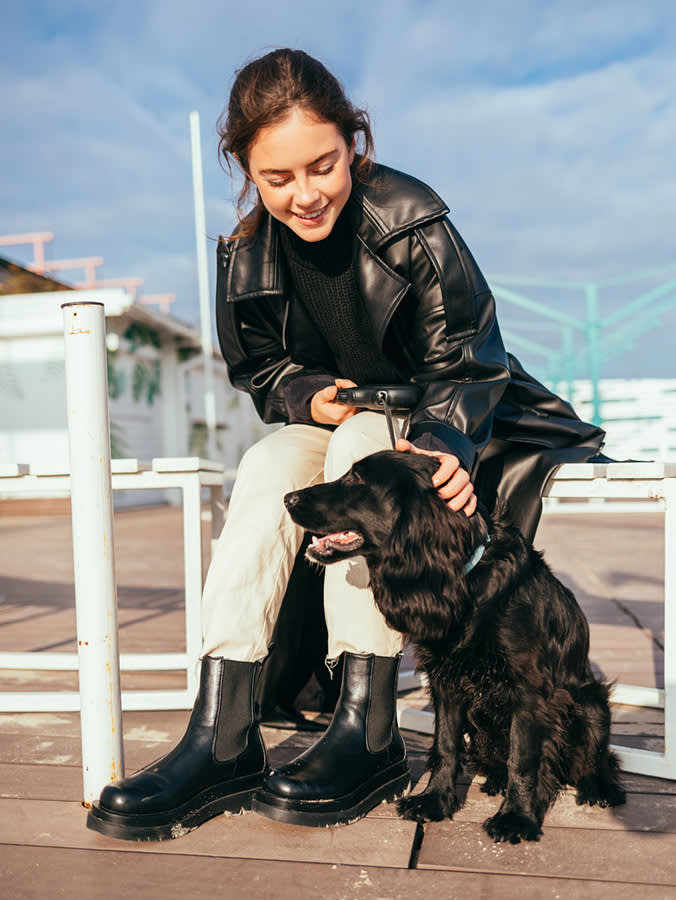 Black puppy on leash lays next to its owner while she sits on bench resting.