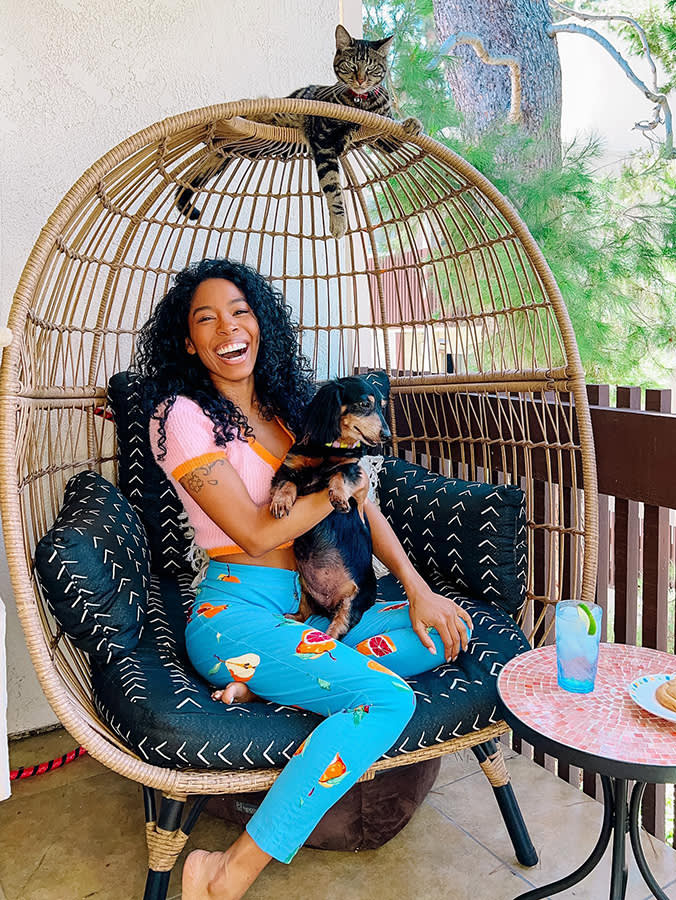 Arisa, a woman with long curly hair wearing bright blue pants and a pink short-sleeve t-shirt sitting in a wicker chair holding her Dachshund dog, Lola, and her cat, Nipsey, laying on top of the chair cage