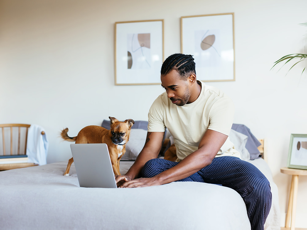 African American man with braids sitting on his bed using his computer with his dog next to him also looking at the computer