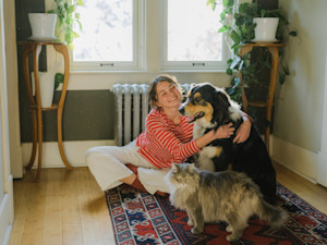 a picture of a young woman sitting on a rug hugging a long haired dog and sitting next to a fluffy cat
