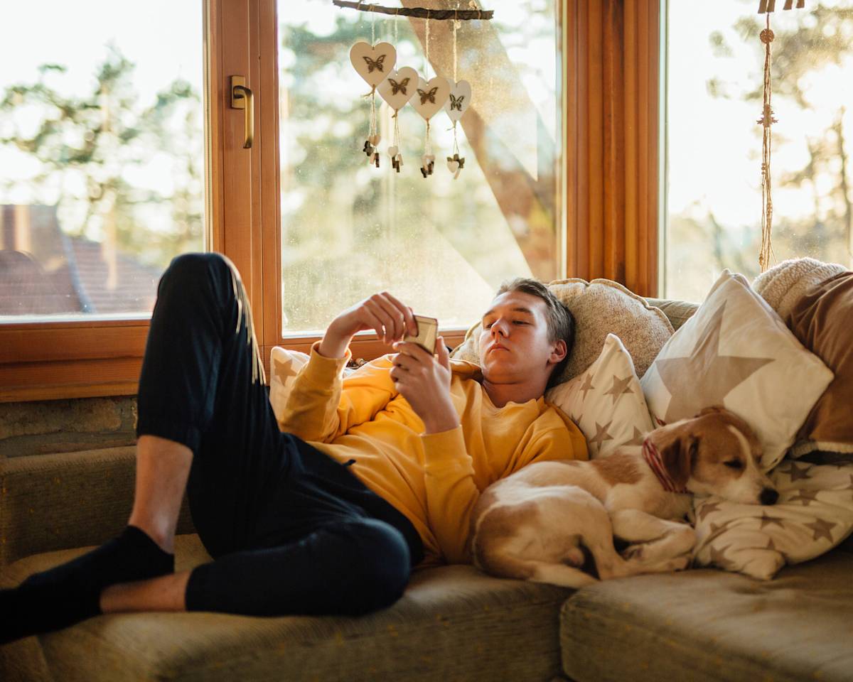 Man in a yellow t-shirt and black pants laying on the couch in an open and airy living room next to his tan and white sleeping dog