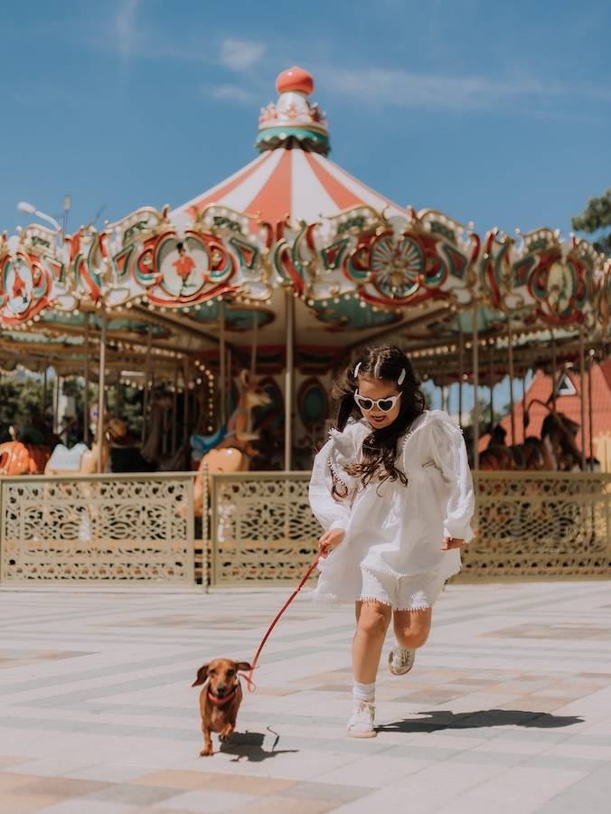 a picture of a little girl in white runs away from a carousel with a daschaund puppy on a lead
