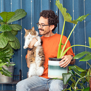 Man holding cat in lap while surrounded by plants.