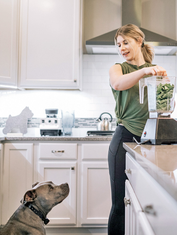 Woman preparing spinach smoothie while gray pit bull dog watches.