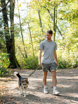 Man wearing grey T-shirt and shorts with trainers walks alongside his dog on the lead in the woods