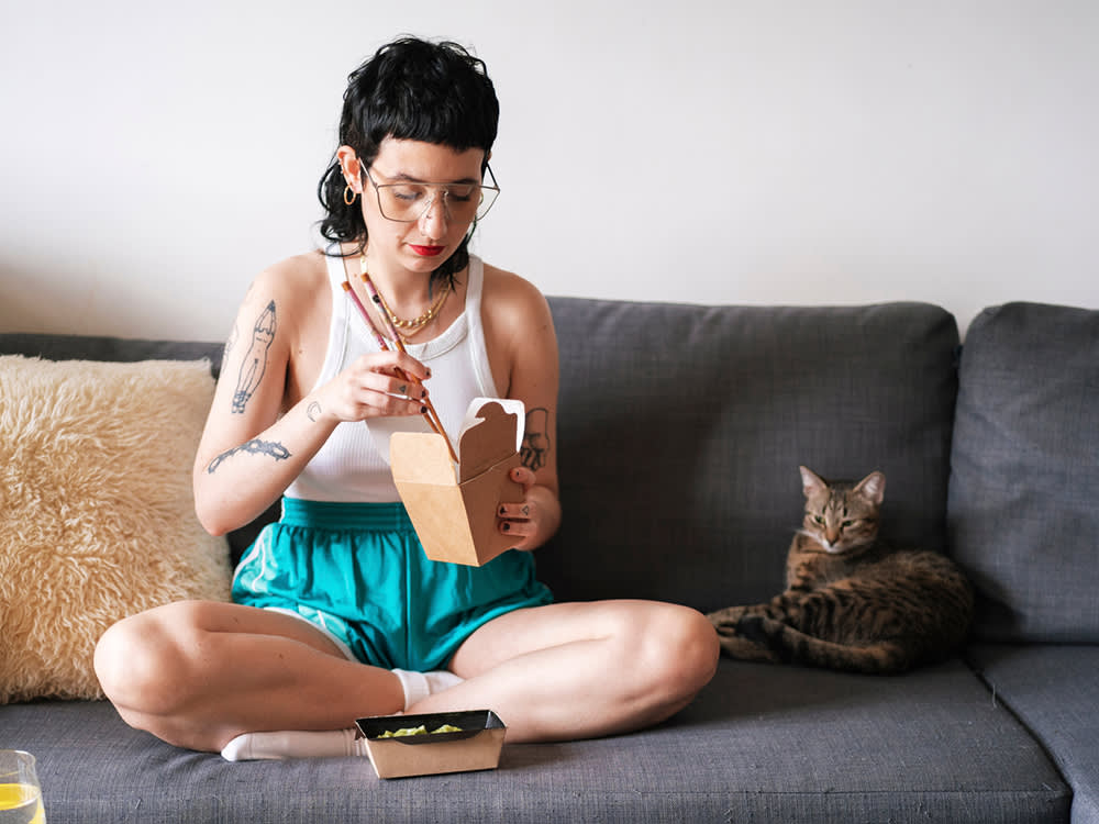 Woman with tattoos eating noodles on the couch with her cat
