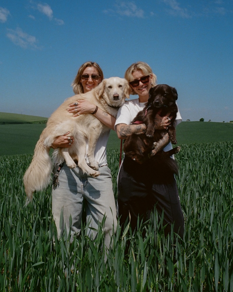 two women in sunglasses with blonde hair hold two large dogs in a field