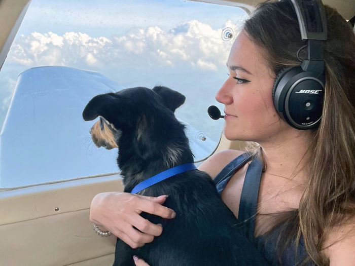 A brunette woman holding a black dog with a blue collar, she is wearing headphones and a microphone attachment  while seated in a plane with the clouds and sky showing through the window 