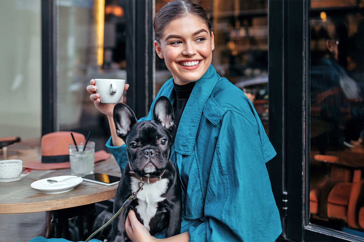 a brunette woman holds a French Bulldog while holding a coffee cup at an outdoor table
