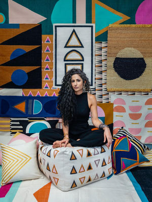 Leah Singh, dressed in all black, sitting cross-legged on a cushion decorated with brightly colored triangles with a vibrant assortment of textiles on the floor and the walls around her, also decorated with bold, geometric designs
