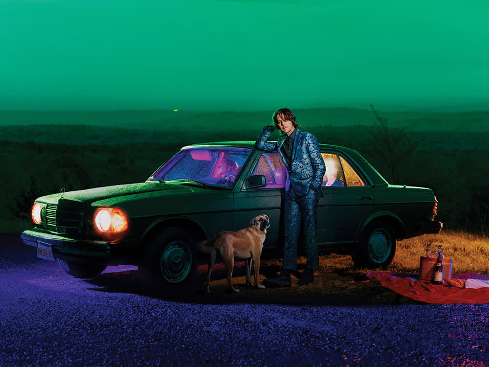Sam Evian and his dog standing in front of a green car set against a staged green landscape on blue gravel