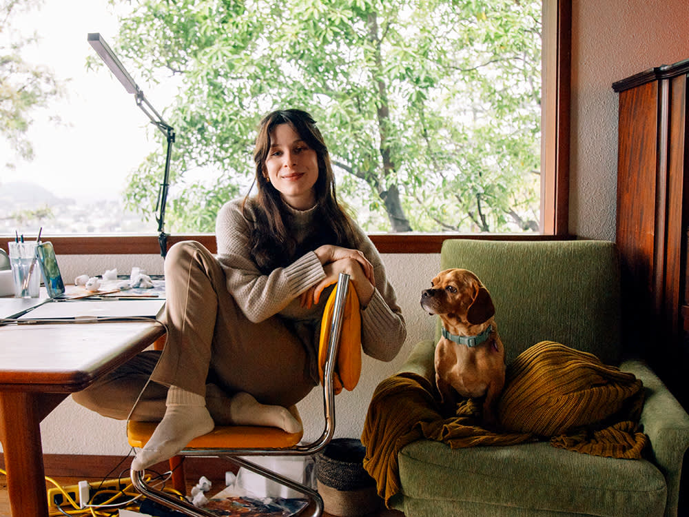 Sasha Spielberg at her desk with her small orange dog beside her on a green chair