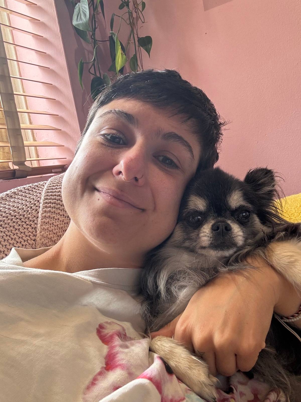 a picture of a person with cropped black hair cuddles their long haired little dog in front of a pink wall