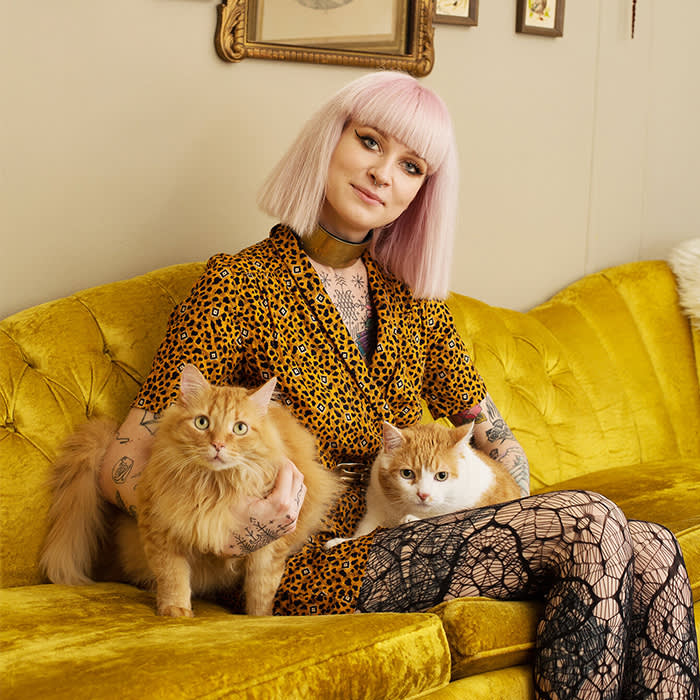 person with pink hair sitting on yellow velvet couch with two orange cats