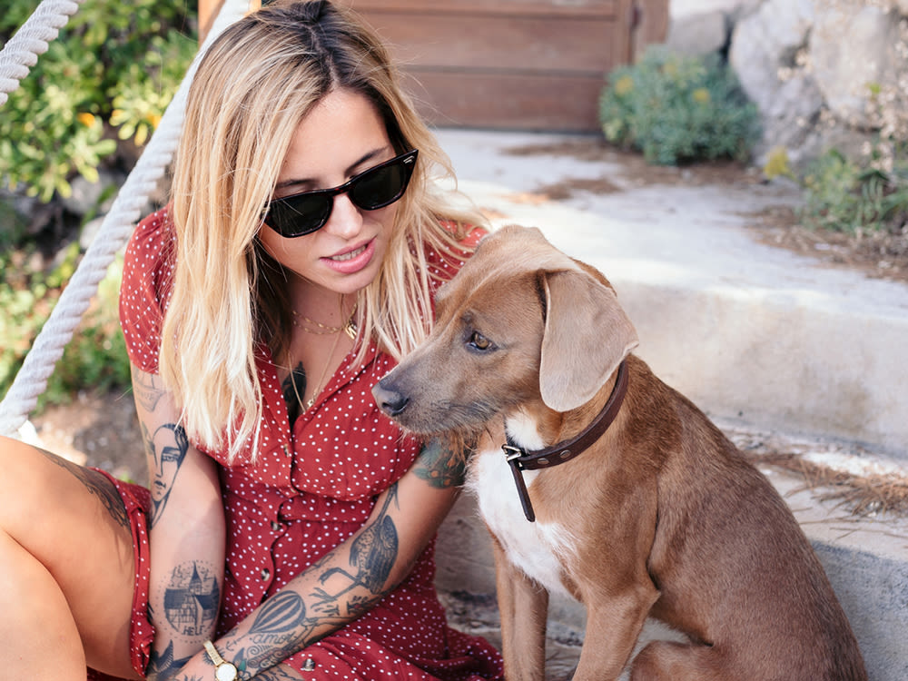 Blonde woman with tattoos in a red dress and sunglasses sits on the front step of her house and talks to her small brown dog 
