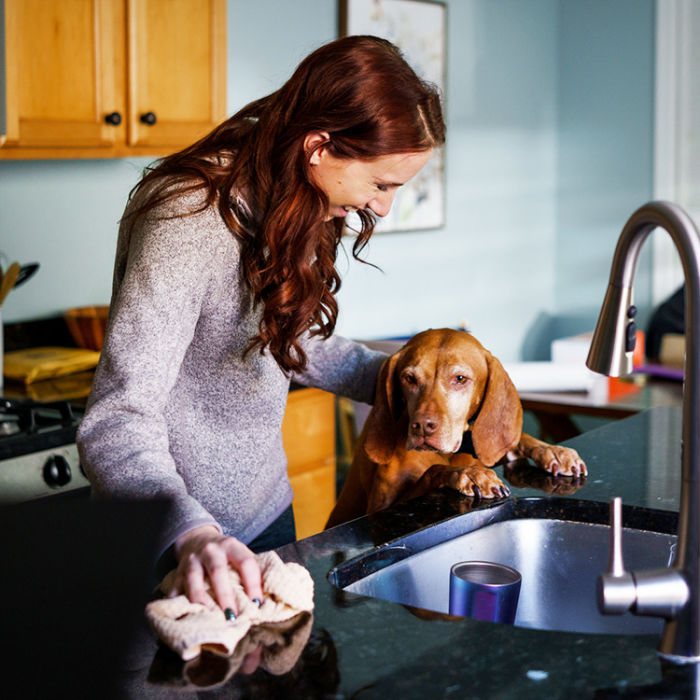 A woman smiling at her dog while cleaning the kitchen counters with a cloth. 