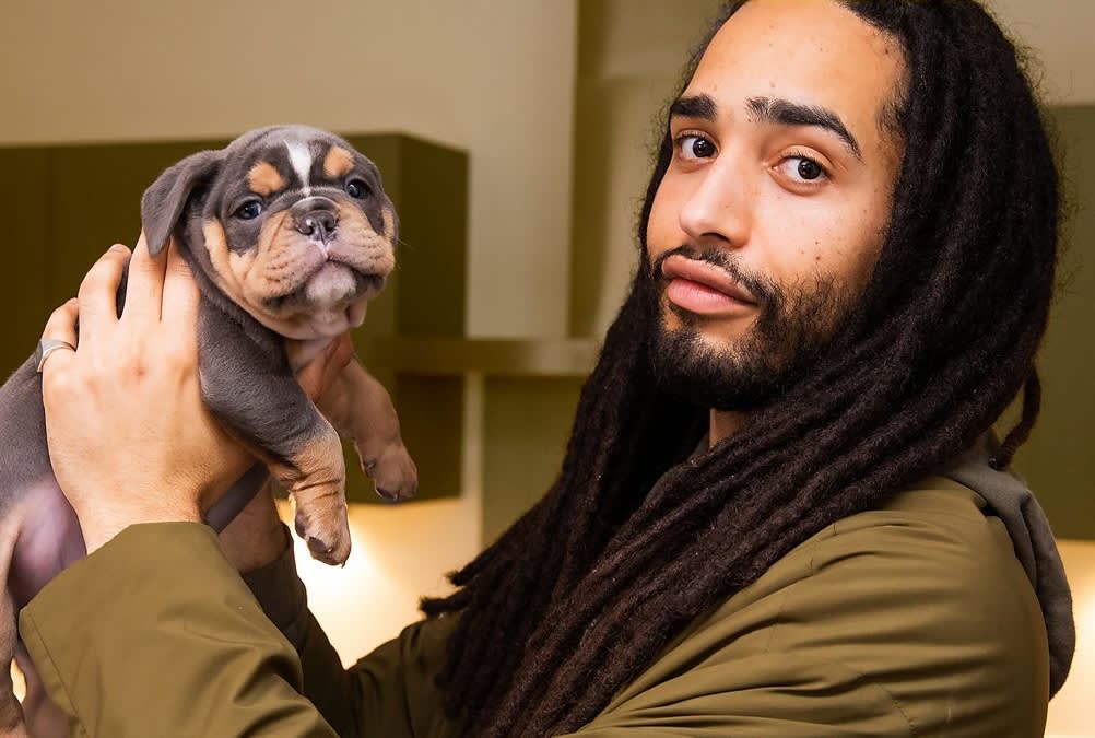 a man with dreadlocks holds up a french bulldog puppy and they both look at the camera