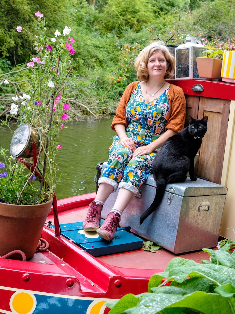 a woman in floral dungarees sits on a narrow boat with a black cat