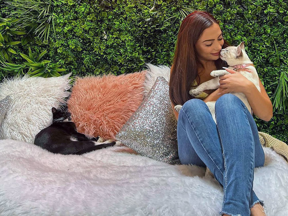 Red-haired woman on white fuzzy bed amidst green bushes holds a beige cat while a black cat stretched out on the bed next to her.
