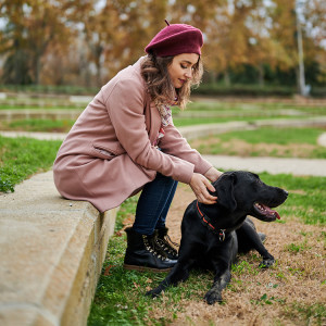 Woman wearing a magenta beret hat and a pink coat sitting on a wall outside and petting her black Labrador dog laying on the ground