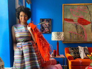 Courtney McLeod wearing a colorful striped dress and a bright orange bag in a blue room with brightly colored and patterned furniture