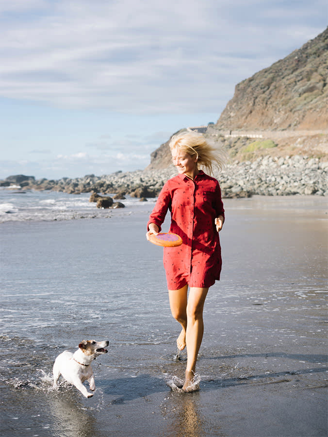 Woman holding frisbee and running on beach with Jack Russell Terrier.
