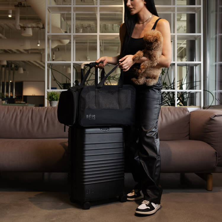 A woman standing next to a suitcase with a dog carrier on top while holding her dog. 