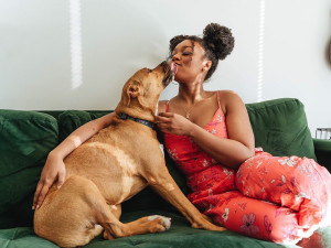 Kassidi Jones sitting on a green velvet couch with her curly hair in two big pom poms and wearing a coral spaghetti strap jumpsuit while her tan dog Gingers Naps licks her face.