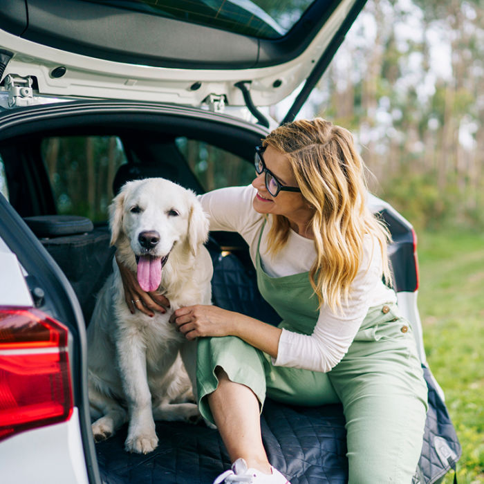 A blonde woman wearing a longsleeved green jumper and sneakers sitting in the open trunk of a SUV car with her arm around her Golden Retriever dog sitting next to her