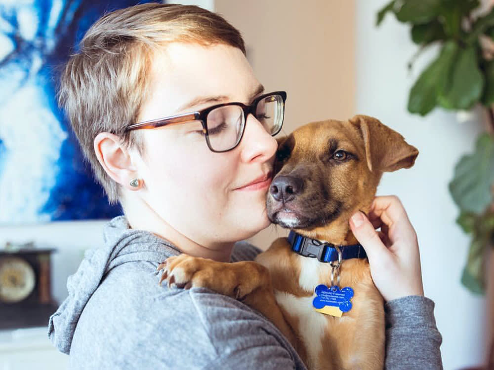 Short-haired blonde woman with glasses holding a mixed brown puppy close to her face affectionately