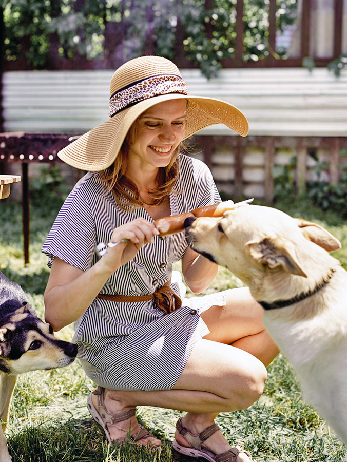 Woman feeding her two dogs hot dogs on a stick outside.