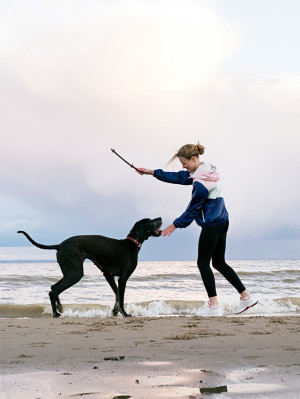 Side view of smiling blonde woman with stick playing with black German Dane pet on beach.