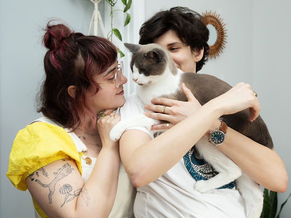 Two woman with short dark hair, one holding a gray and white cat while the other leans against the woman holding the cat, to look at the cat