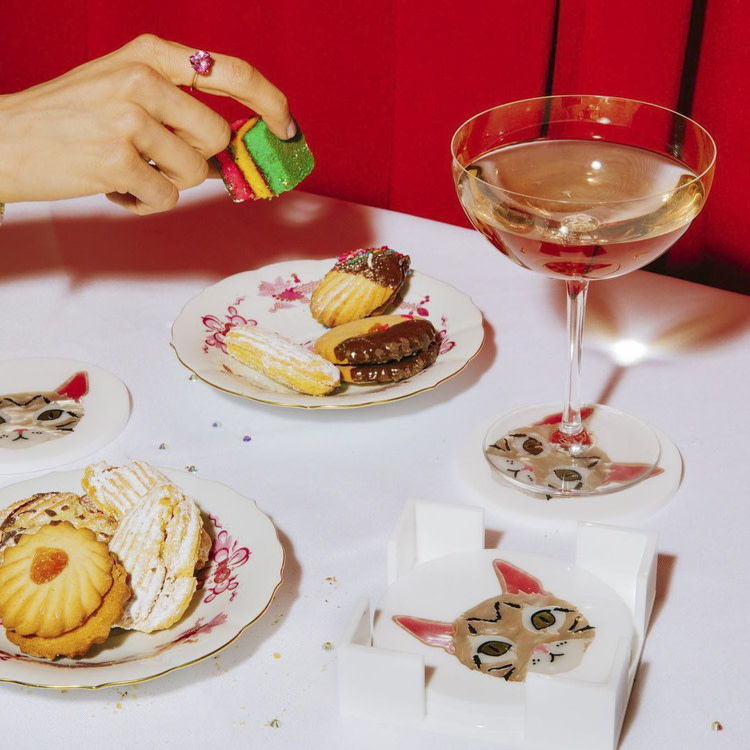 A still life photo of Edie Parker cat coasters, plates with small deserts, and a champagne glass. 