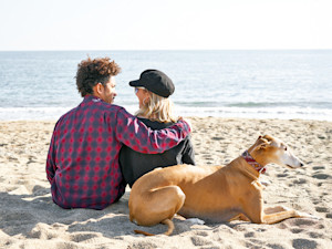 Couple sitting on the beach with a dog behind them.