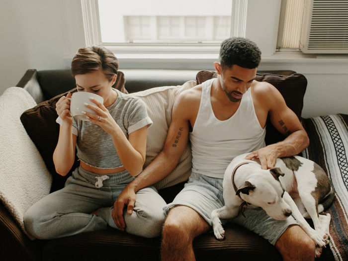 A woman sitting on a couch sipping a cup of coffee and a man sitting next to her with a dog on his lap. 