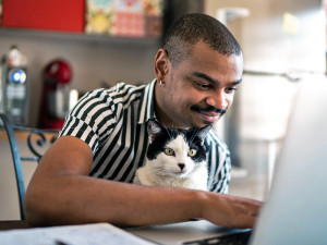 A man sitting at his kitchen table using a laptop with a black and white cat on his lap.