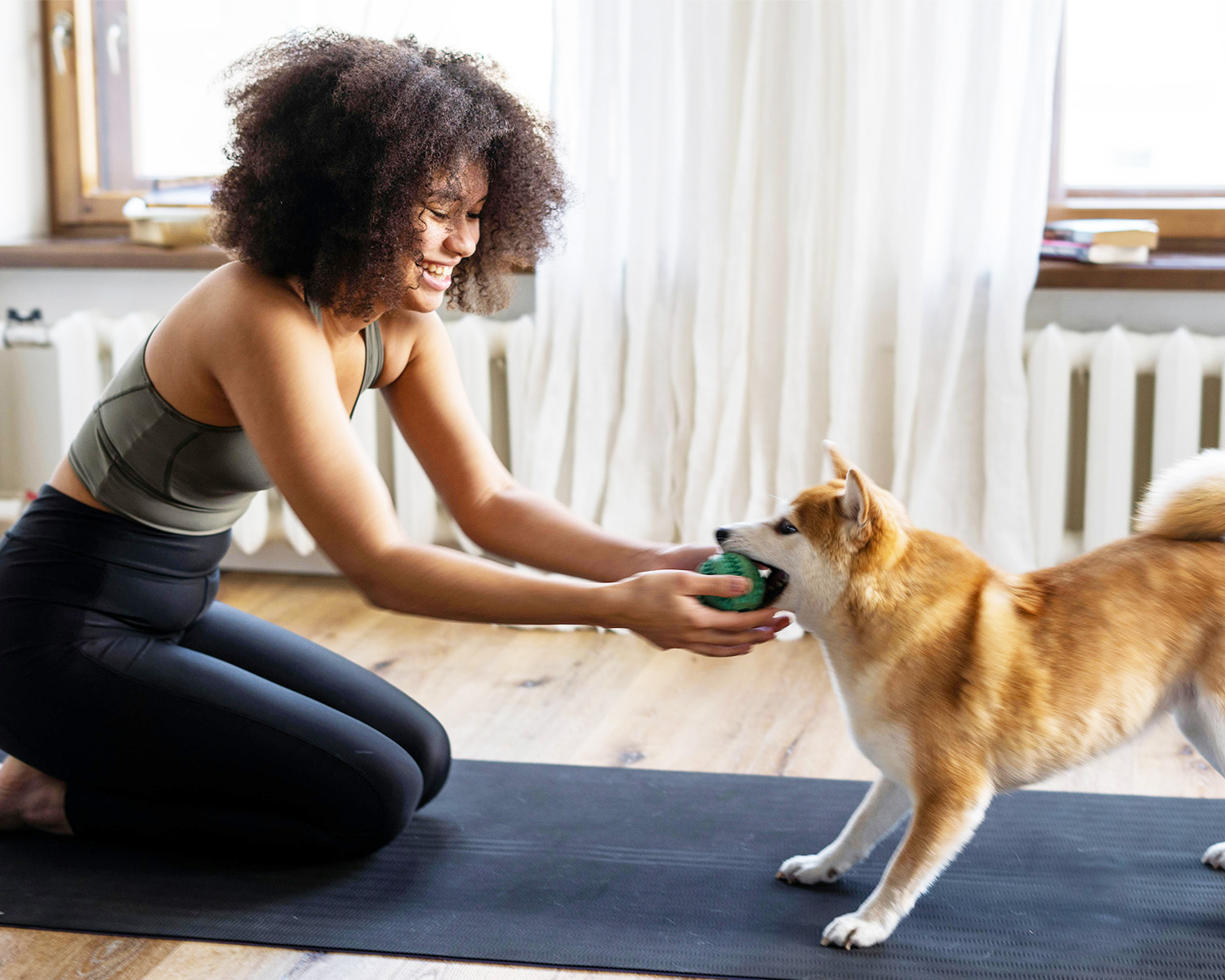 a Black woman in yoga kit plays with a Shiba inu with a green ball
