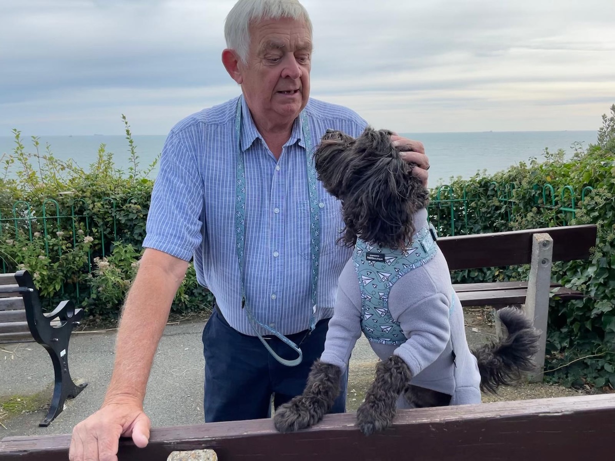 an older man pets a dog at a bench with the sea behind them