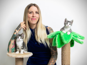 The Wildest's cover star Hannah Shaw smiling at the camera behind a cat tower with two kittens sitting on top of it