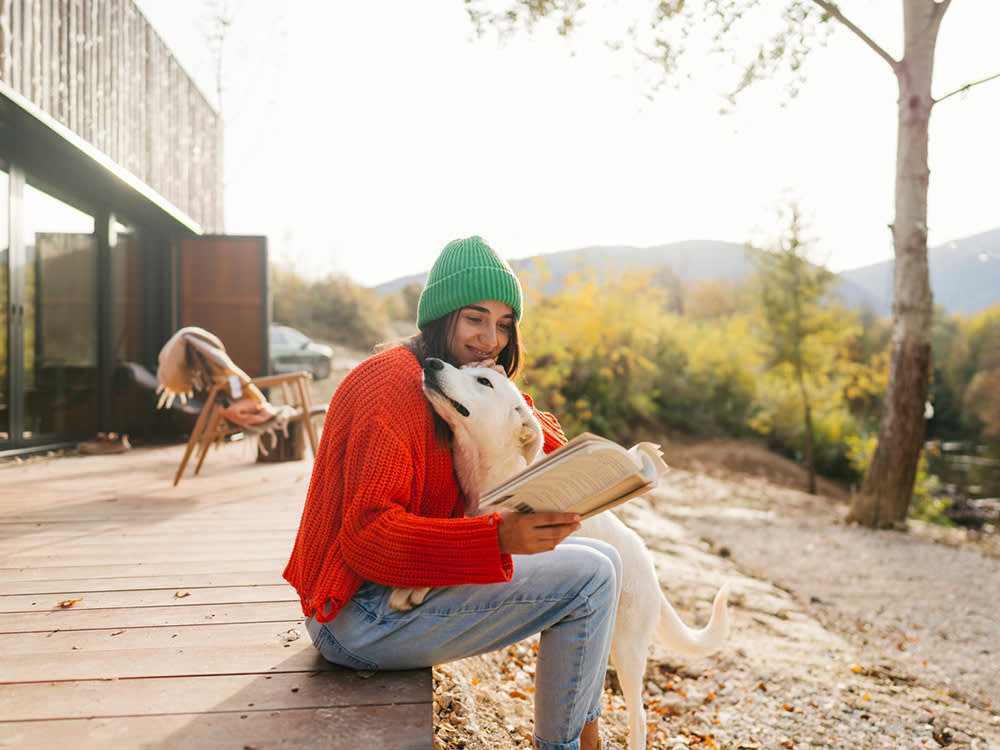 a woman in a red sweater and green hat hugs a dog outside while reading a book
