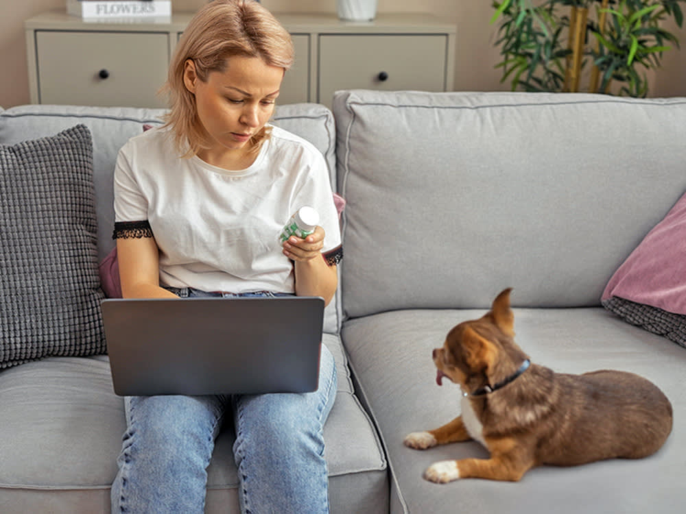 Woman looking at pill bottle while small dog sits on couch next to her.