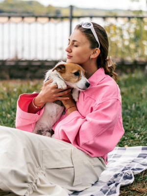 A young positive woman in a pink jacket and sunglasses hugging friendly Jack Russell Terrier dog, enjoying happy moments together while walking in the park on a summer day.
