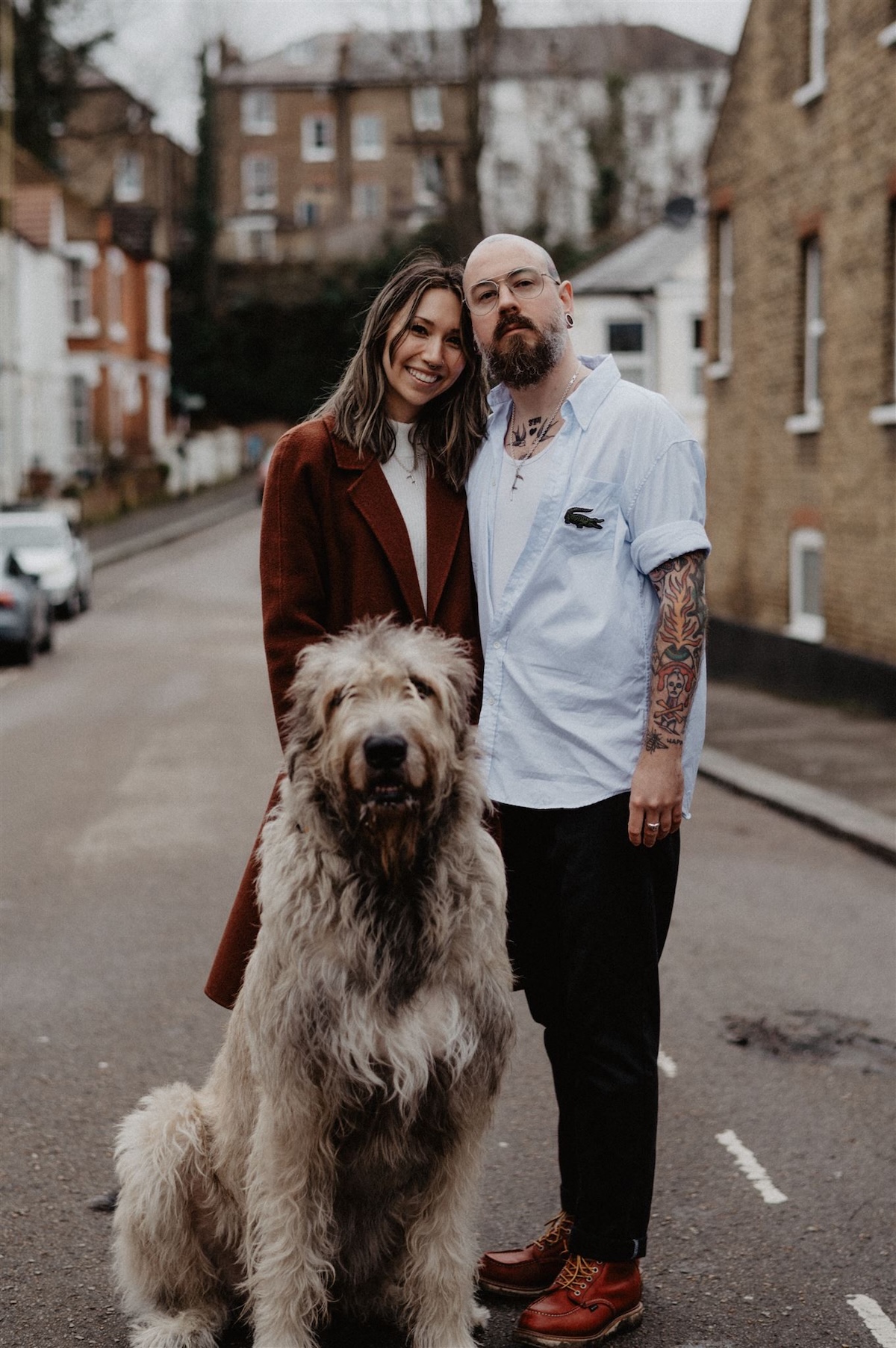a well dressed alternative couple stand on the street with the gigantic shaggy dog