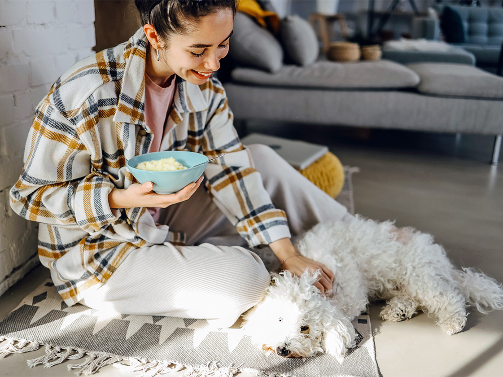 Woman eating mashed potatoes and petting her small white dog.