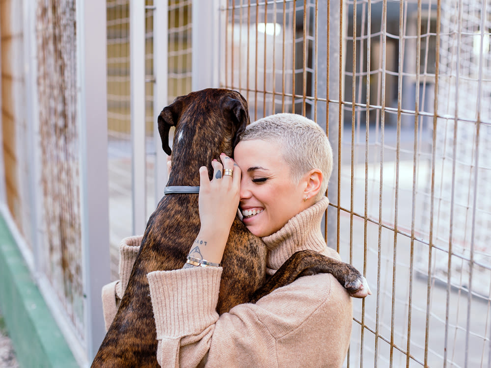 A woman with tattoos with close shaved blonde hair wearing a tan sweater hugging her merle coat dog outside of a dog shelter